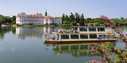 MS Schärding mit Personen an Bord im Inn mit Kloster Neuhaus im Hintergrund und dem Link  zu den Angeboten